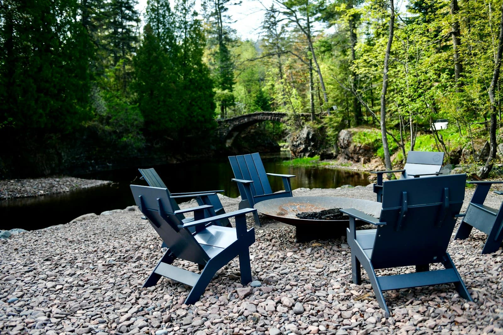 Adirondack chairs around a fire pit on a gravel patio at the edge of the woods