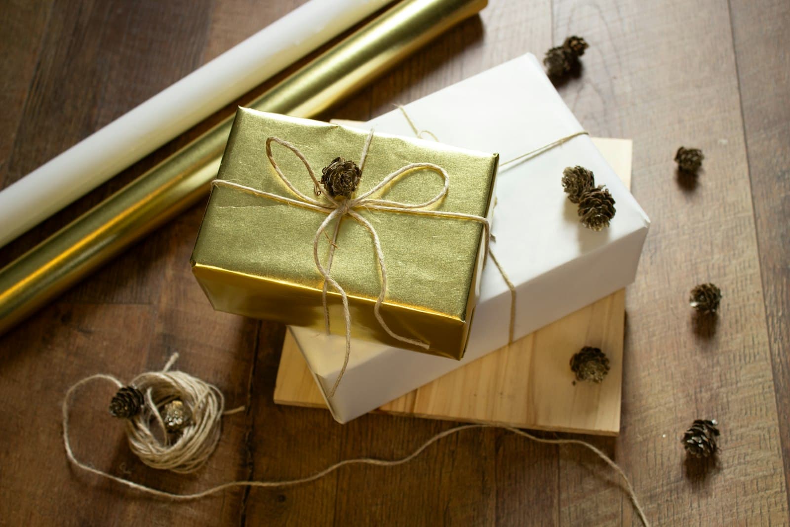 Rolls of gold and white wrapping paper beside a gold-wrapped gift tied with twine, stacked on a white-wrapped box, with a loose coil of twine and small pinecones