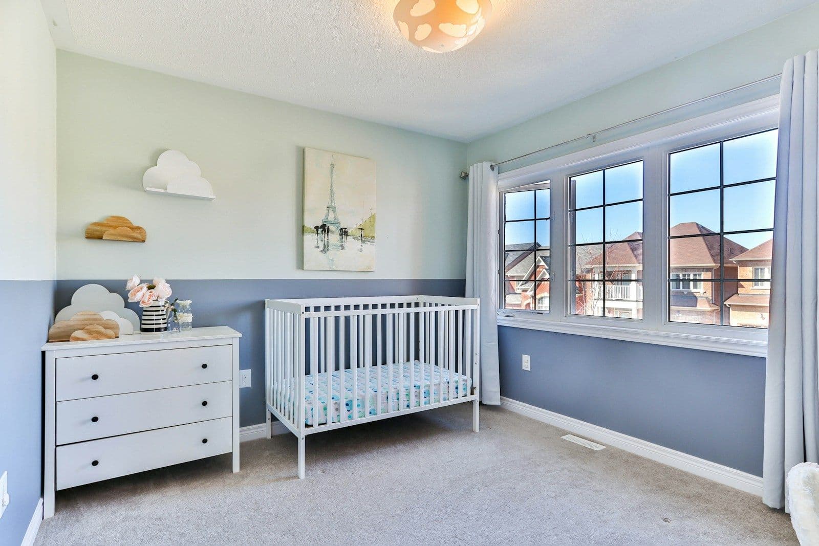 A bare white crib beside a white dresser in a blue and white nursery with curtained windows