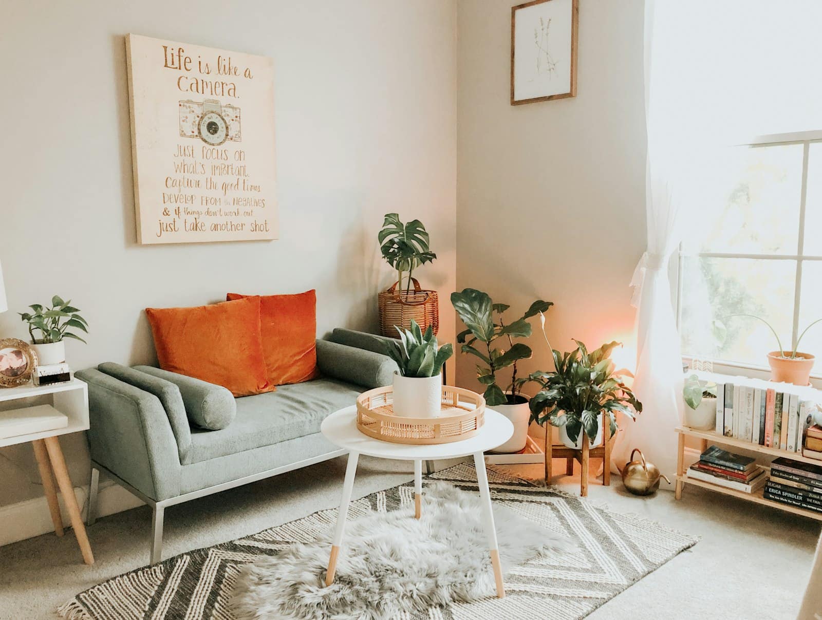 A small, bright living room with a sage loveseat, orange pillows, and plants