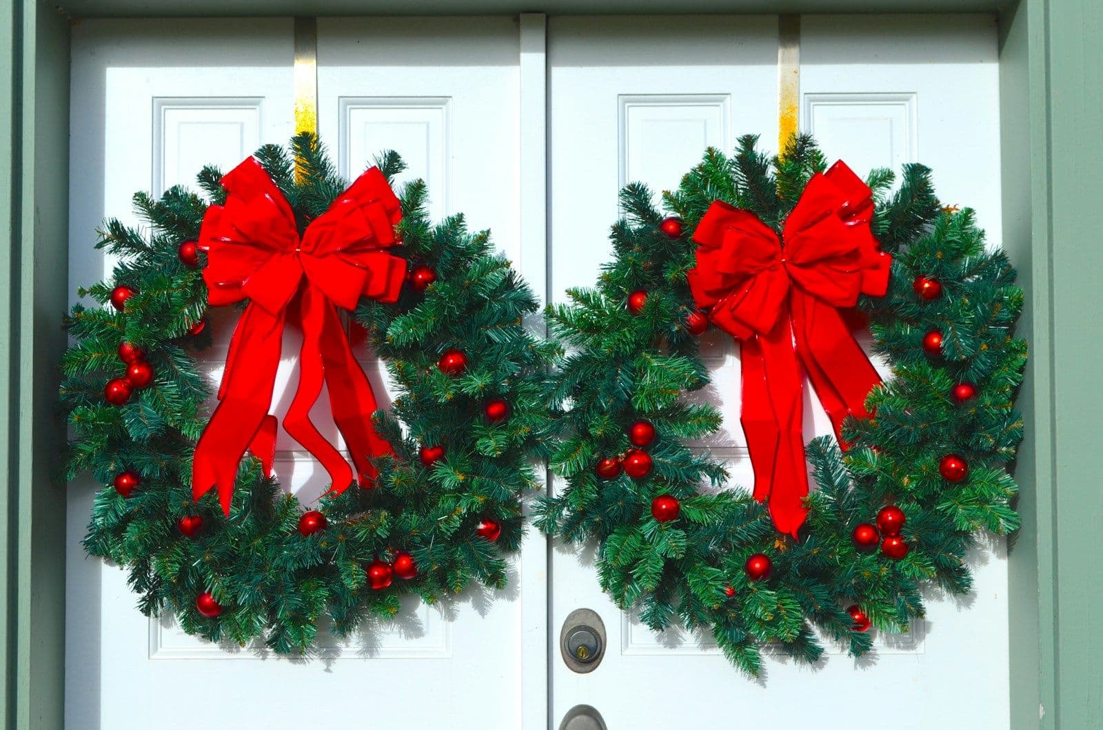 Two evergreen wreaths with red bows and berries hanging on a white double front door