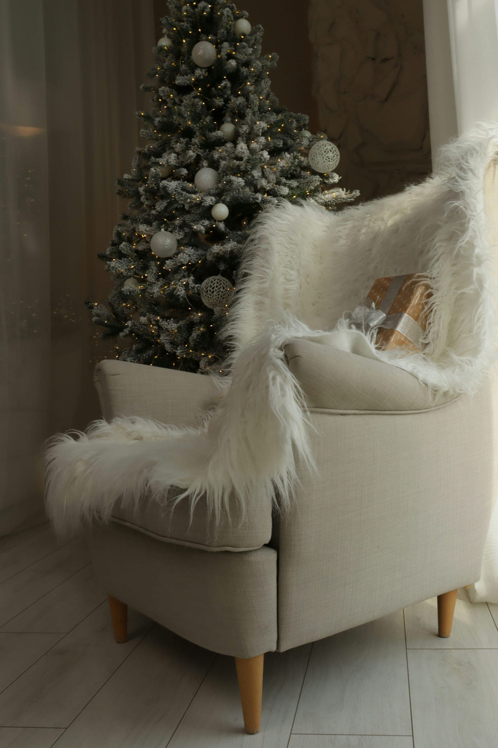 A cream armchair draped with a white faux fur throw and a wrapped gift, beside a snow-dusted Christmas tree with white ornaments