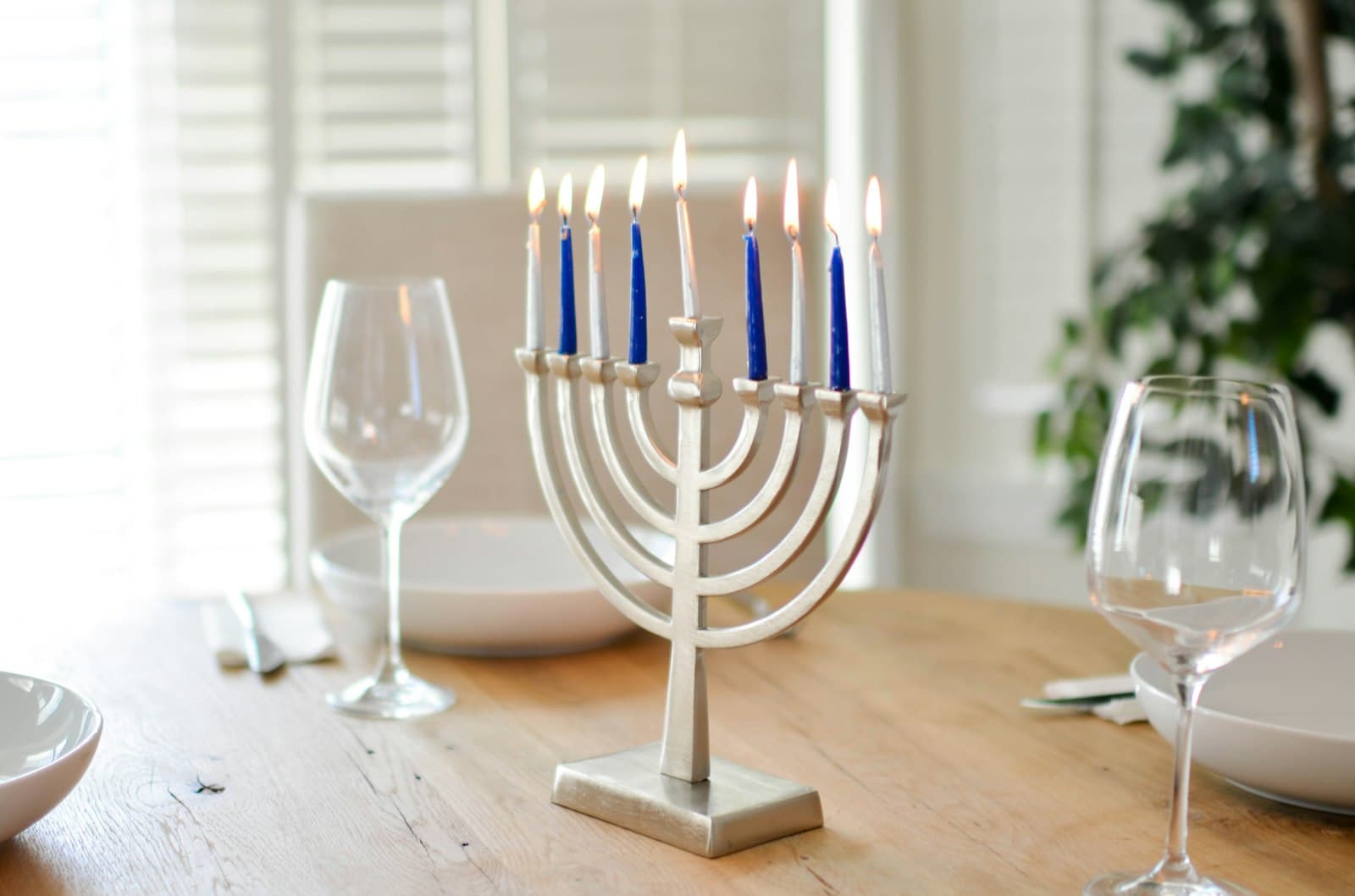 A brushed-silver menorah with all nine blue and white candles lit, on a wood dining table set with white plates and wine glasses