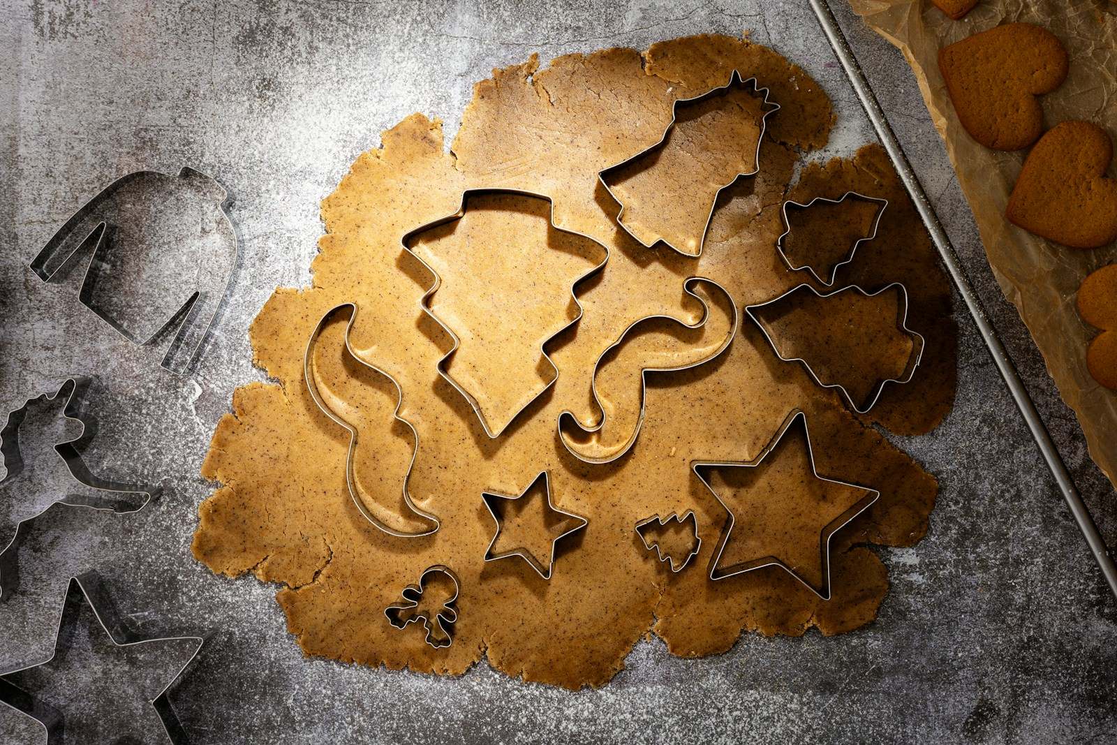 Star, tree, bell and gingerbread-man cutters pressed into rolled-out gingerbread dough on a floured counter, beside a baking sheet of heart-shaped cookies