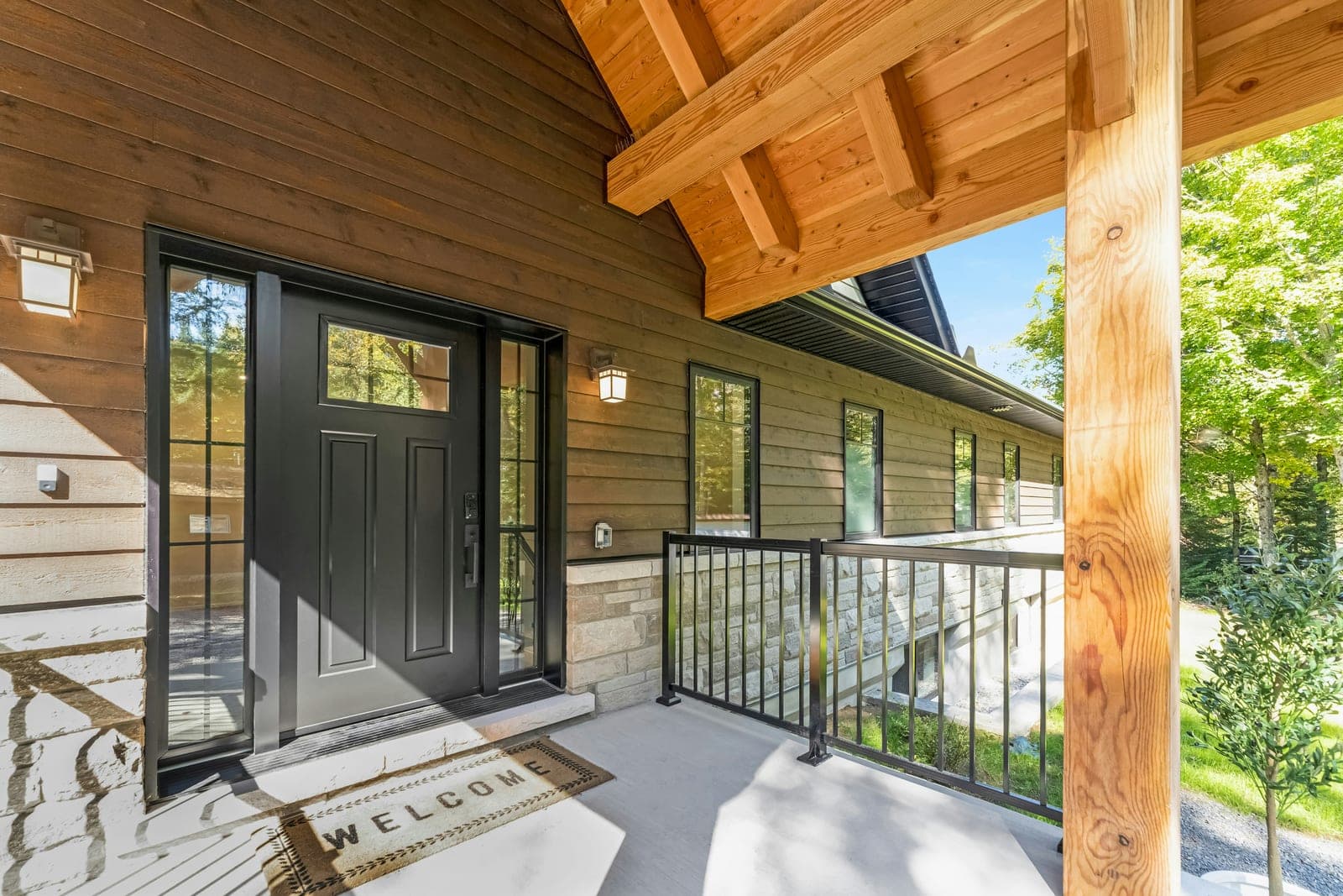 A black front door with sidelights and a welcome mat on the covered porch of a newly built wood-sided house