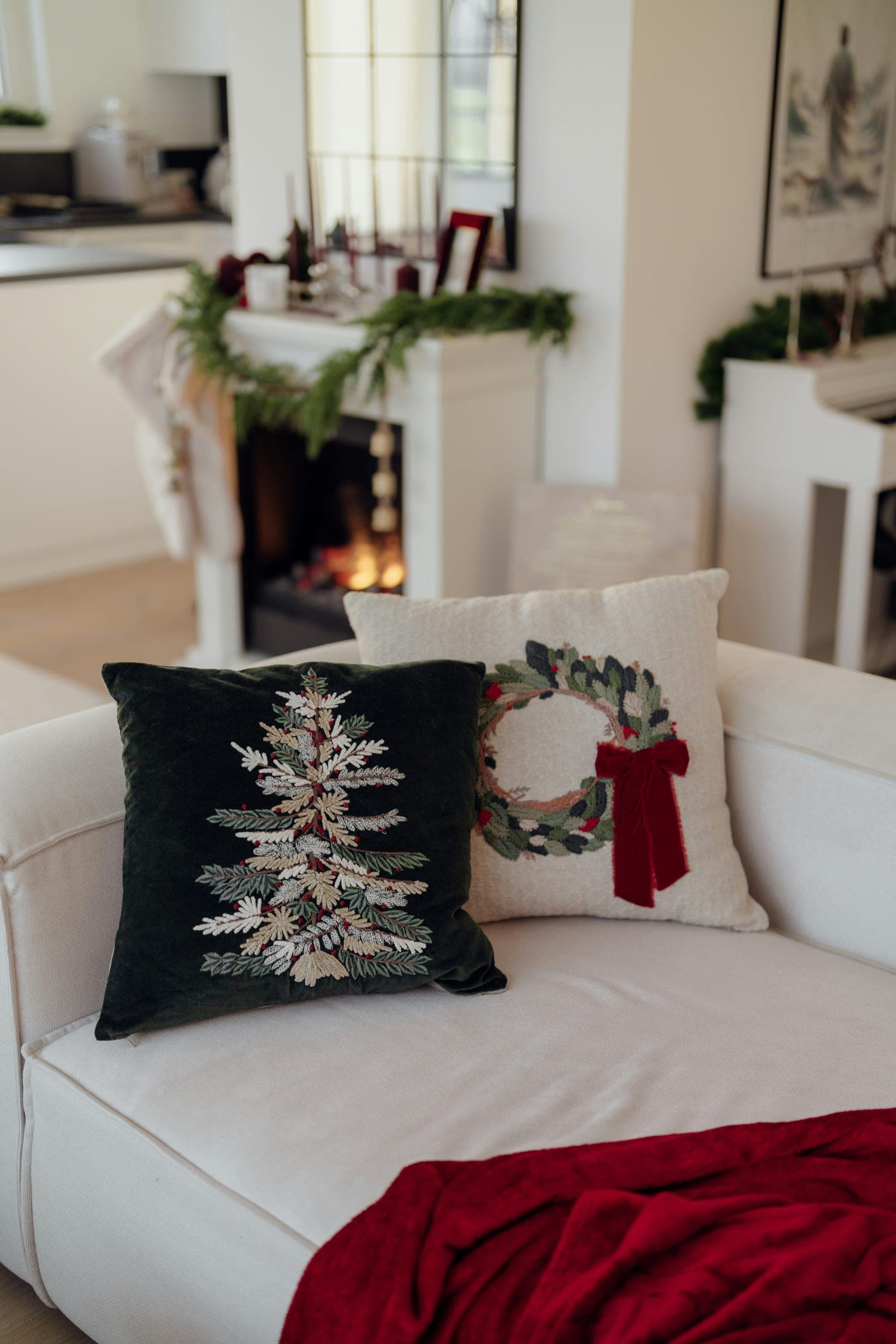 A white sofa with a dark green velvet pillow embroidered with a tree and a cream pillow with a wreath, a red throw across the seat, and a lit fireplace with a garland on the mantel behind it